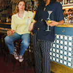 A woman sitting on a barstool holding a margarita wearing a Tombolo shirt next to a man standing holding a martini, wearing a Tombolo shirt with an embroidered martini on it