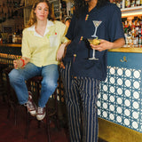 A woman sitting on a barstool holding a margarita wearing a Tombolo shirt next to a man standing holding a martini, wearing a Tombolo shirt with an embroidered martini on it