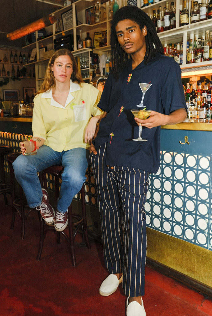 A woman sitting on a barstool holding a margarita wearing a Tombolo shirt next to a man standing holding a martini, wearing a Tombolo shirt with an embroidered martini on it