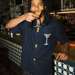 Image of model standing at the bar, sipping on a dirty martini, wearing the 'Extra Dirty' ~ Relaxed Fit Shirt and our Striped Easy Going Pants. 