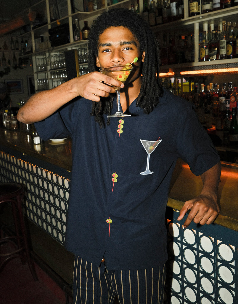 Image of model standing at the bar, sipping on a dirty martini, wearing the 'Extra Dirty' ~ Relaxed Fit Shirt and our Striped Easy Going Pants. 