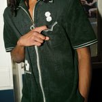 Man wearing a dark green terry cloth short-sleeve shirt stands indoors with one hand casually pointing toward his chest pocket. His body is angled slightly, with the other hand tucked in his pocket. The shirt features embroidered dice above the pocket and playing cards in a lower pocket, with white piping and a visible zipper running down the front.
