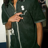Man wearing a dark green terry cloth short-sleeve shirt stands indoors with one hand casually pointing toward his chest pocket. His body is angled slightly, with the other hand tucked in his pocket. The shirt features embroidered dice above the pocket and playing cards in a lower pocket, with white piping and a visible zipper running down the front.