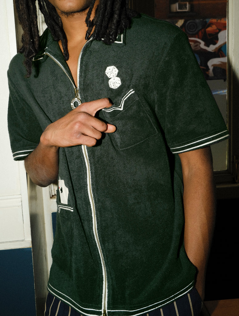Man wearing a dark green terry cloth short-sleeve shirt stands indoors with one hand casually pointing toward his chest pocket. His body is angled slightly, with the other hand tucked in his pocket. The shirt features embroidered dice above the pocket and playing cards in a lower pocket, with white piping and a visible zipper running down the front.