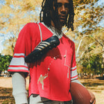 A person wearing a red mesh football-style crop top with cowrie shells forming a football graphic, layered over a gray long-sleeve shirt and paired with gray athletic shorts, holding a football with cleats draped over one shoulder.