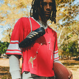 A person wearing a red mesh football-style crop top with cowrie shells forming a football graphic, layered over a gray long-sleeve shirt and paired with gray athletic shorts, holding a football with cleats draped over one shoulder.