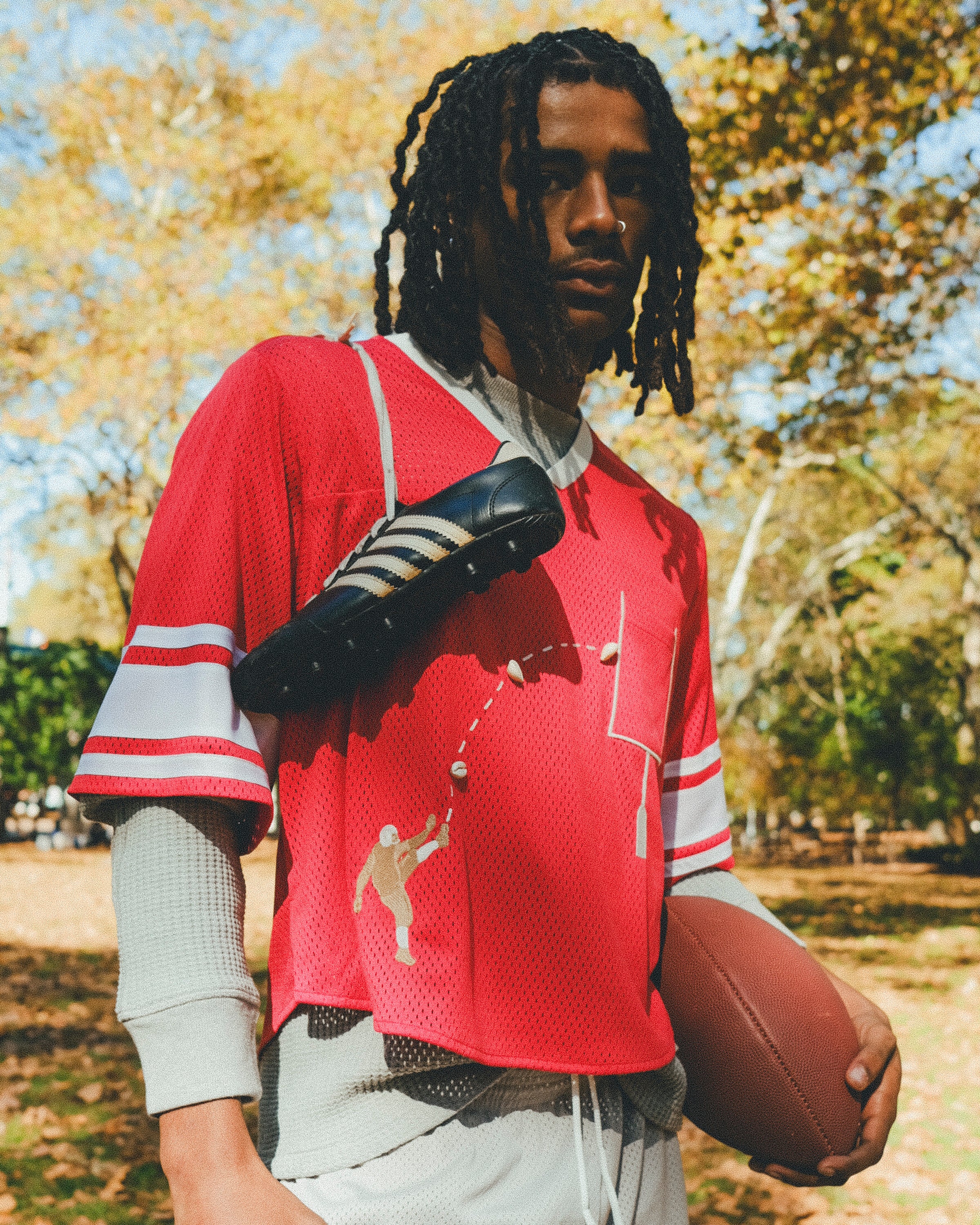A person wearing a red mesh football-style crop top with cowrie shells forming a football graphic, layered over a gray long-sleeve shirt and paired with gray athletic shorts, holding a football with cleats draped over one shoulder.