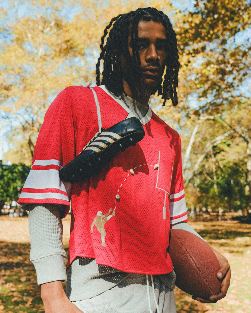 A person wearing a red mesh football-style crop top with cowrie shells forming a football graphic, layered over a gray long-sleeve shirt and paired with gray athletic shorts, holding a football with cleats draped over one shoulder.