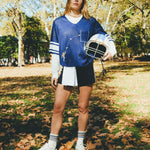 A woman wearing a navy blue football-style crop shirt, layered over a white long-sleeve shirt and paired with navy shorts, striped socks, and white sneakers while holding a white helmet.