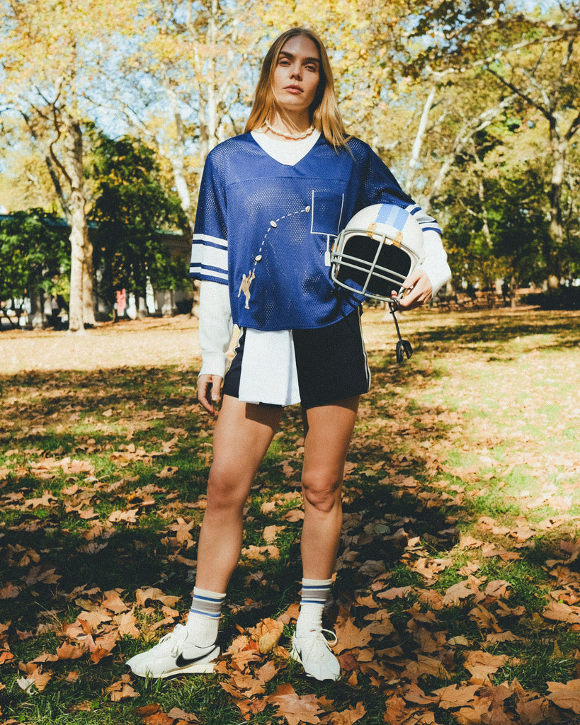 A woman wearing a navy blue football-style crop shirt, layered over a white long-sleeve shirt and paired with navy shorts, striped socks, and white sneakers while holding a white helmet.