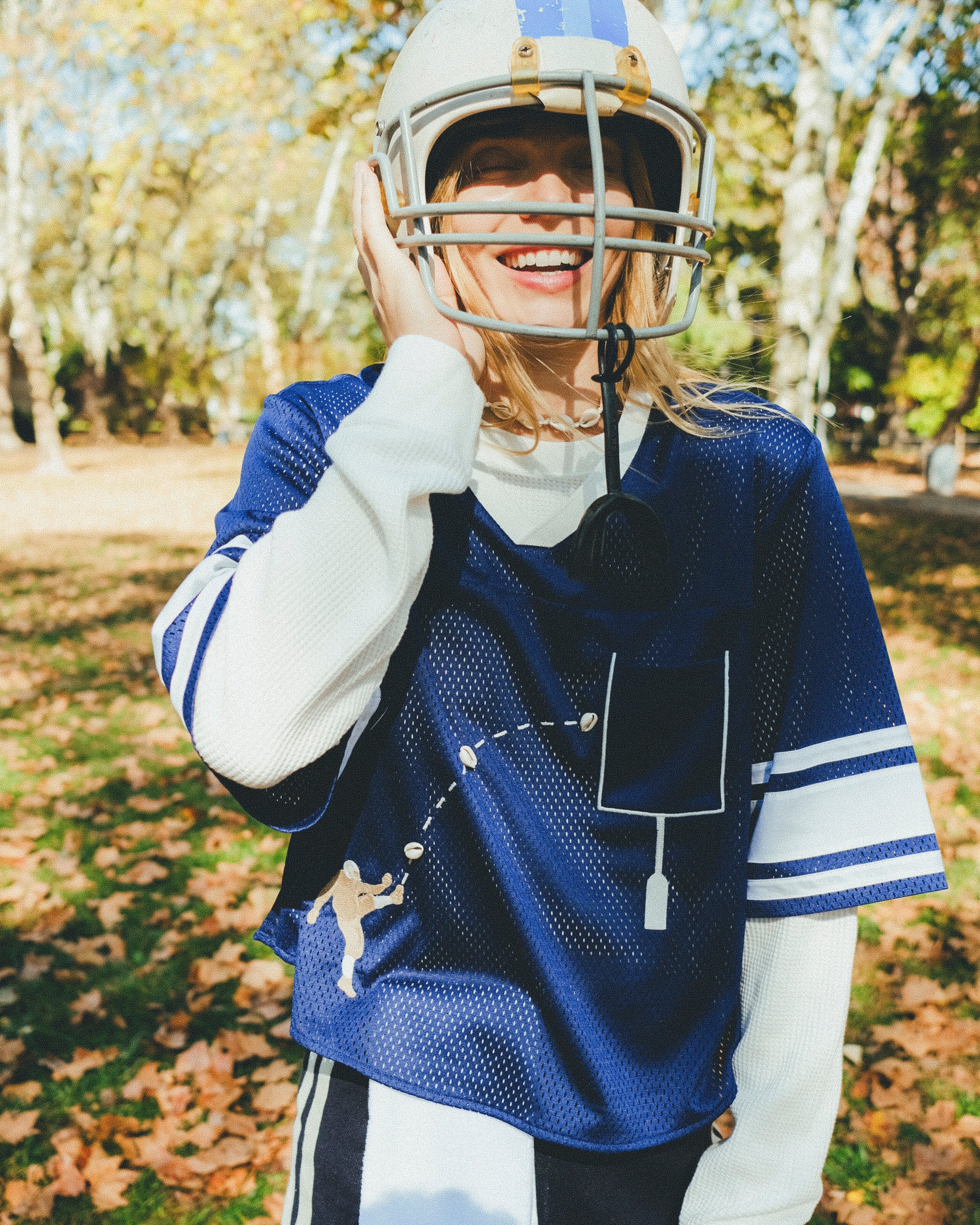 Woman wearing the navy blue mesh football jersey over a white long sleeve shirt. She is laughing and wearing a football helmet. 