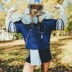 A woman in a blue sports jersey, a helmet and shoulder pads, standing outdoors with arms raised.