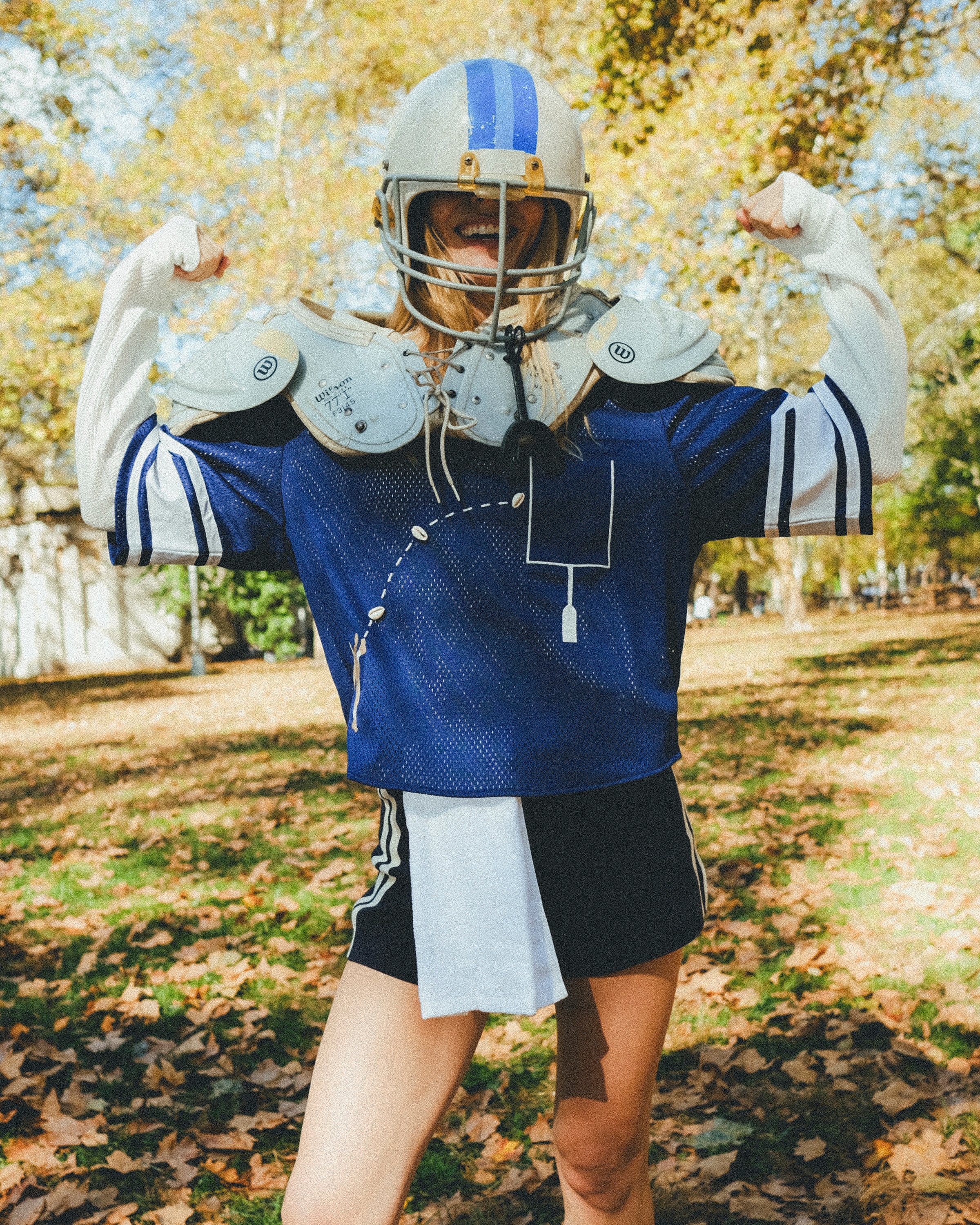 A woman in a blue sports jersey, a helmet and shoulder pads, standing outdoors with arms raised.
