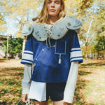 Woman wearing mesh blue sports jersey with football shoulders pads and carrying a helmet in her hand.