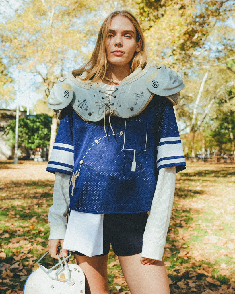 Woman wearing mesh blue sports jersey with football shoulders pads and carrying a helmet in her hand.