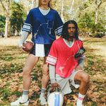 Two people wearing football-style mesh tops with cowrie shells forming a football graphic. One wears a navy blue version with layered long sleeves and shorts, holding a football. The other wears a red version with layered sleeves and shorts, holding a helmet.