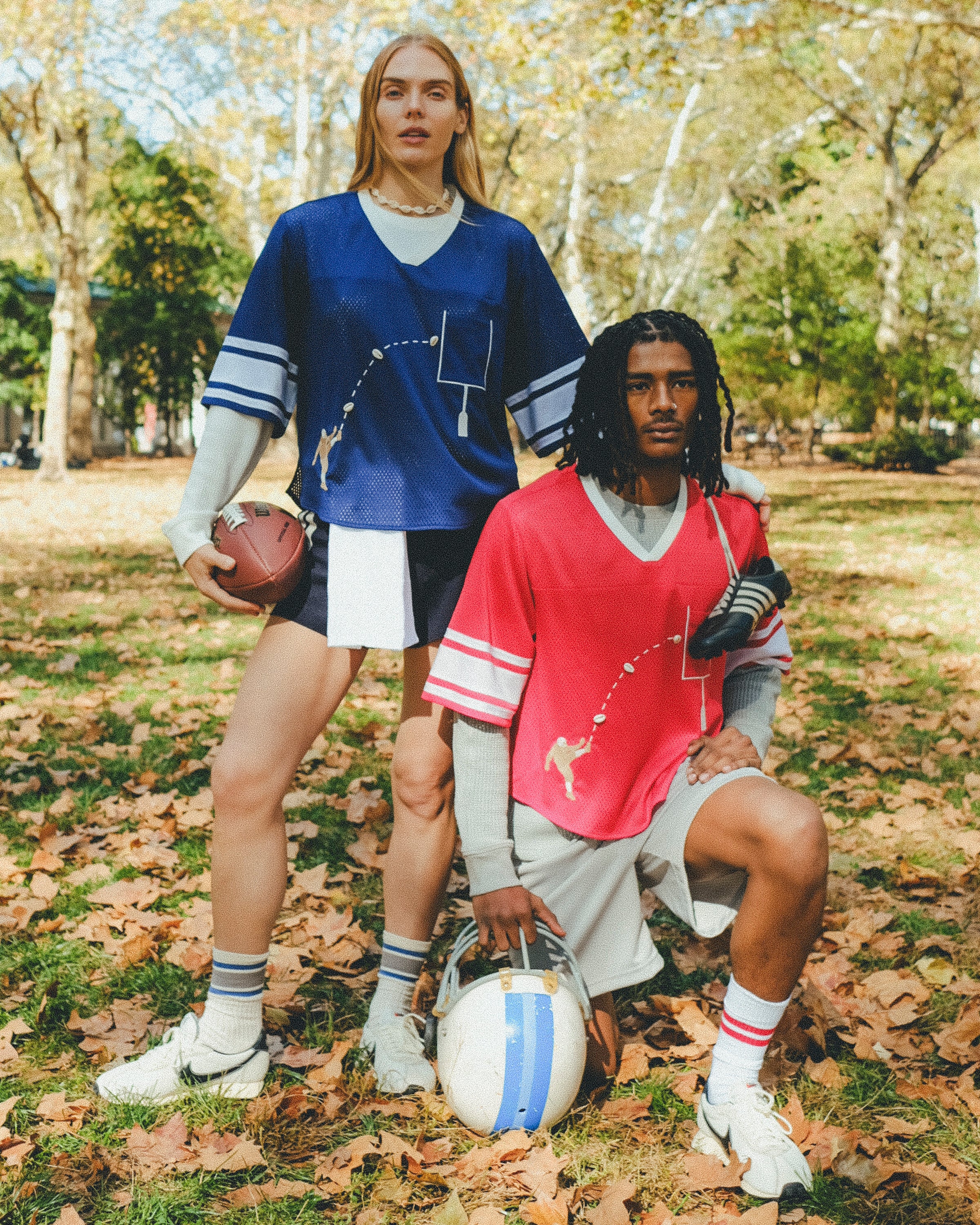 Two people wearing football-style mesh tops with cowrie shells forming a football graphic. One wears a navy blue version with layered long sleeves and shorts, holding a football. The other wears a red version with layered sleeves and shorts, holding a helmet.