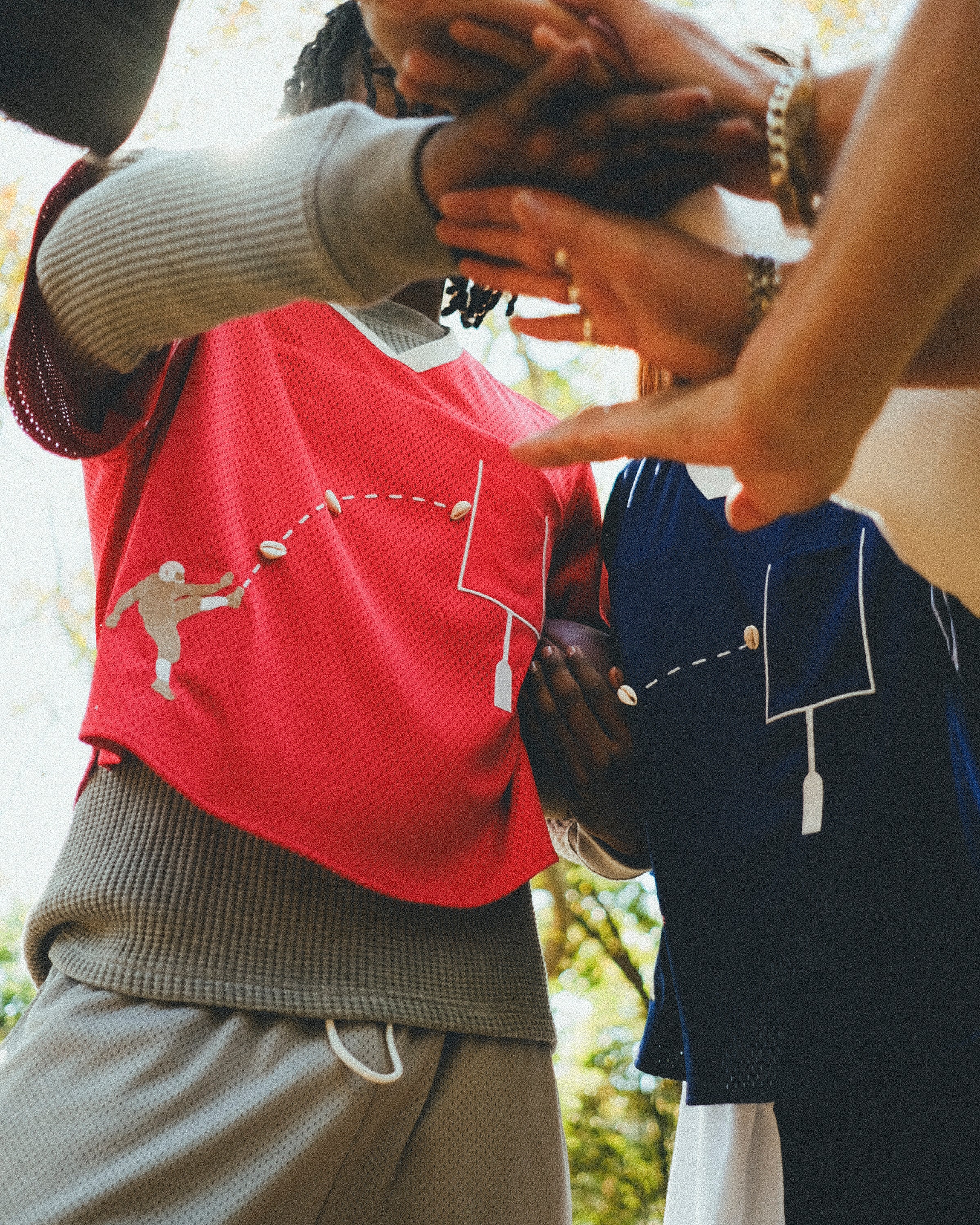 Two people wearing red and navy mesh football-style tops with cowrie shell football graphics, joining hands in a team huddle.