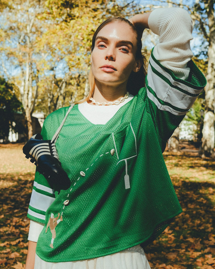 A woman wearing a green mesh football-style crop top with cowrie shells forming a football graphic, layered over a white long-sleeve shirt.