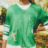 Close-up of a woman wearing a green mesh football-style crop top with cowrie shells forming a football graphic, layered over a white long-sleeve shirt.