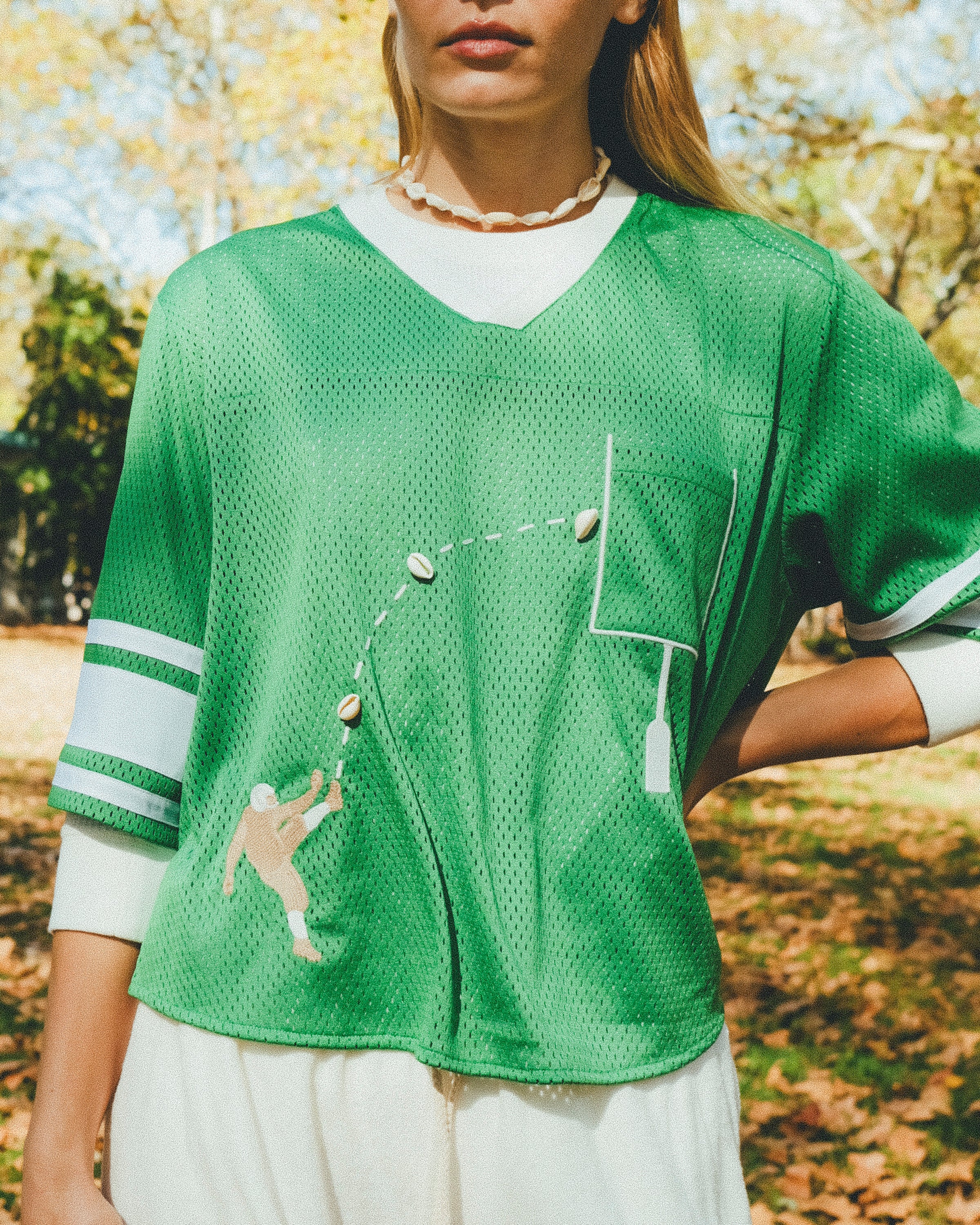 Close-up of a woman wearing a green mesh football-style crop top with cowrie shells forming a football graphic, layered over a white long-sleeve shirt.