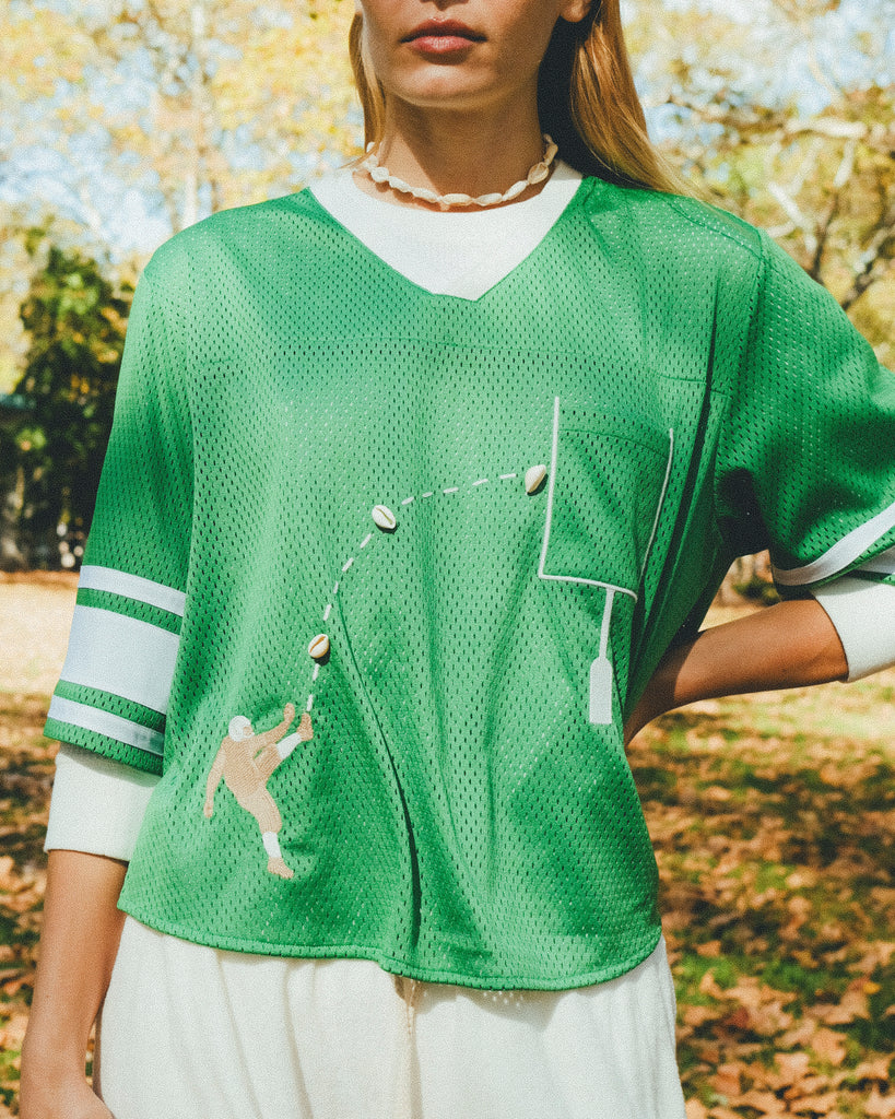 Close-up of a woman wearing a green mesh football-style crop top with cowrie shells forming a football graphic, layered over a white long-sleeve shirt.