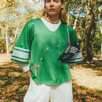A woman wearing a green mesh football-style crop top with cowrie shells forming a football graphic, layered over a white long-sleeve shirt and paired with white shorts, with cleats draped over one shoulder.