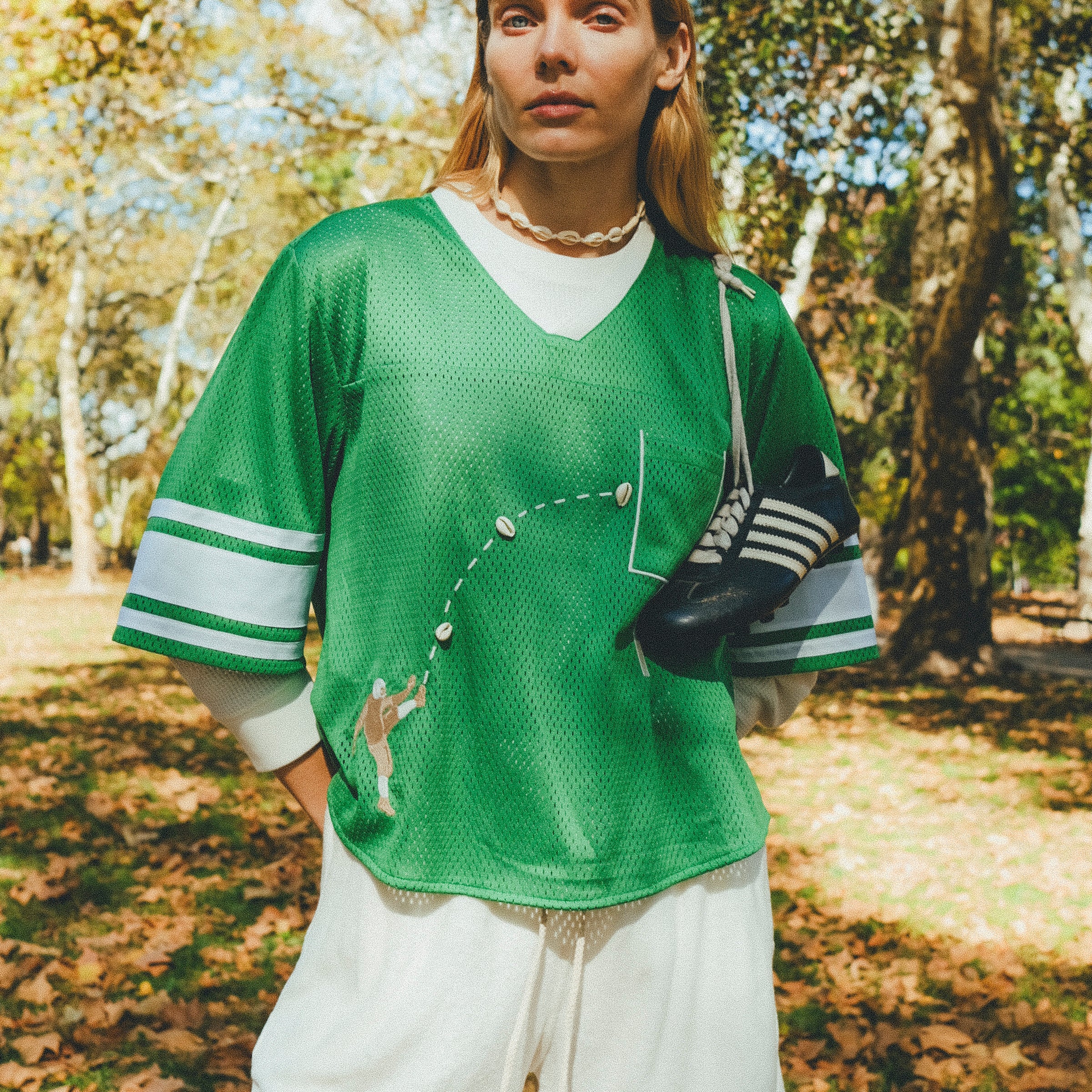 A woman wearing a green mesh football-style crop top with cowrie shells forming a football graphic, layered over a white long-sleeve shirt and paired with white shorts, with cleats draped over one shoulder.