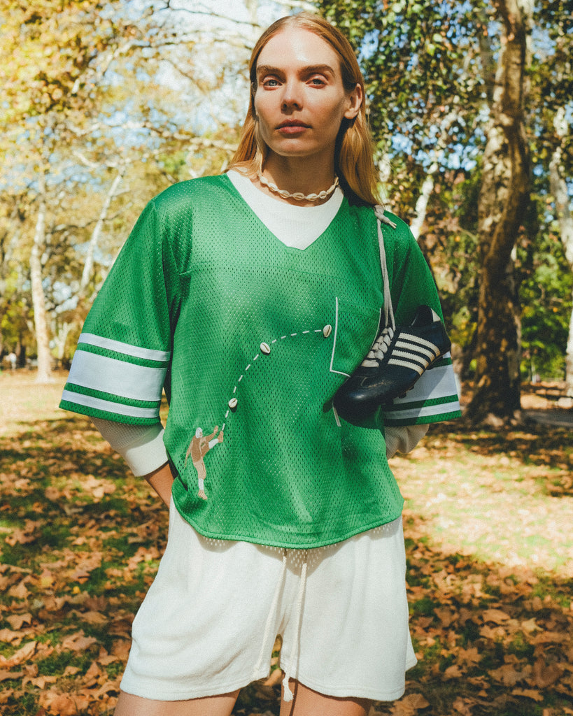 A woman wearing a green mesh football-style crop top with cowrie shells forming a football graphic, layered over a white long-sleeve shirt and paired with white shorts, with cleats draped over one shoulder.