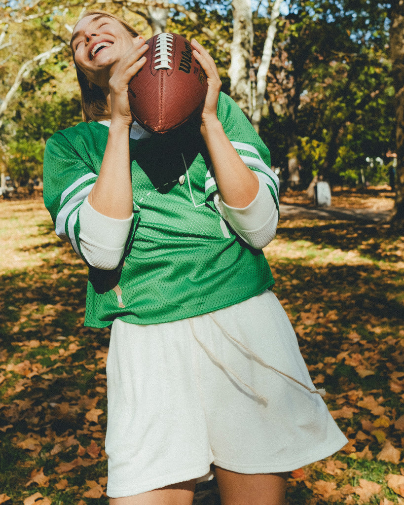 Woman in green mesh sports jersey and white shorts holding a football outdoors.
