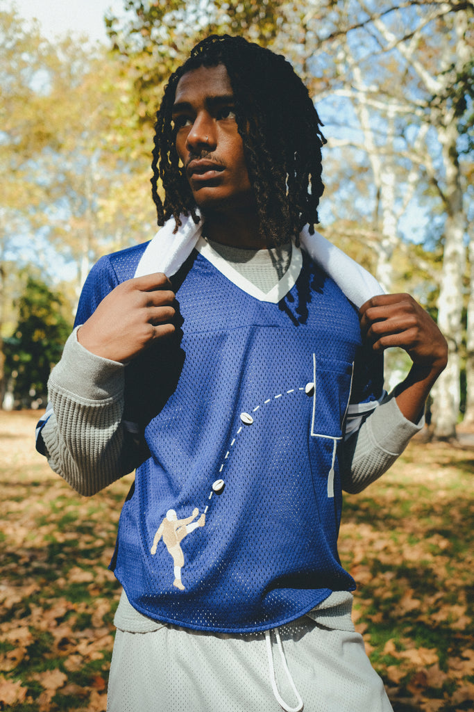 A man wearing the blue mesh football jersey, with a gray long sleeve layered underneath. He is carrying a towel around his neck. 