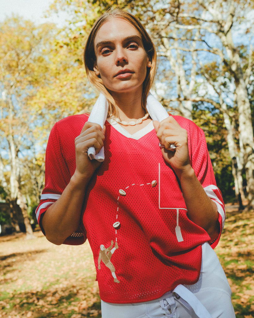 Woman wearing a red mesh football jersey and standing in a park, wearing a towel around her neck.
