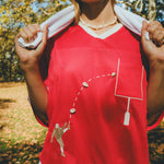Close-up of a red mesh football-style top with cowrie shells forming a football graphic and an outlined pocket, worn over a white shirt with a towel draped around the neck.
