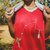 Close-up of a red mesh football-style top with cowrie shells forming a football graphic and an outlined pocket, worn over a white shirt with a towel draped around the neck.