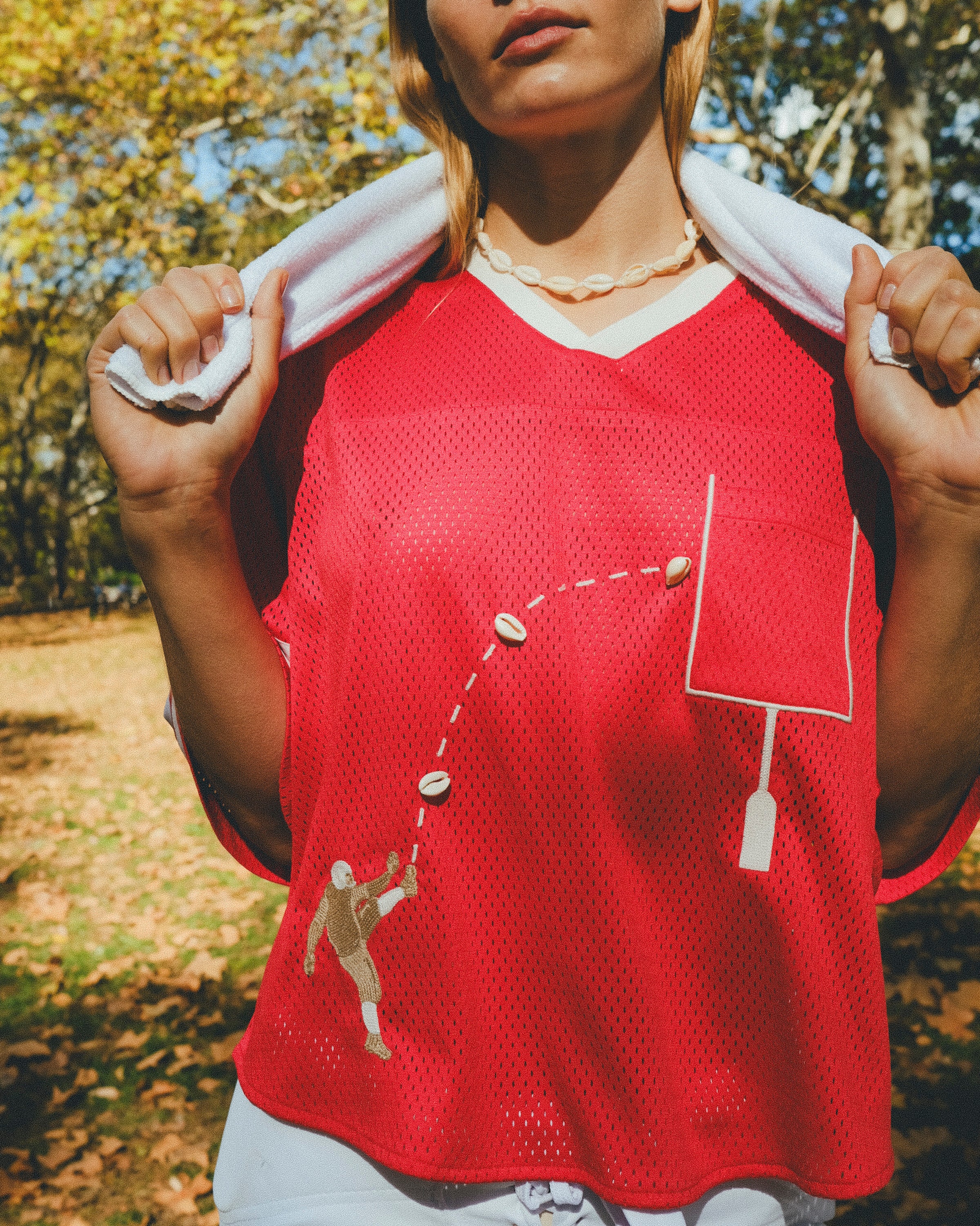 Close-up of a red mesh football-style top with cowrie shells forming a football graphic and an outlined pocket, worn over a white shirt with a towel draped around the neck.