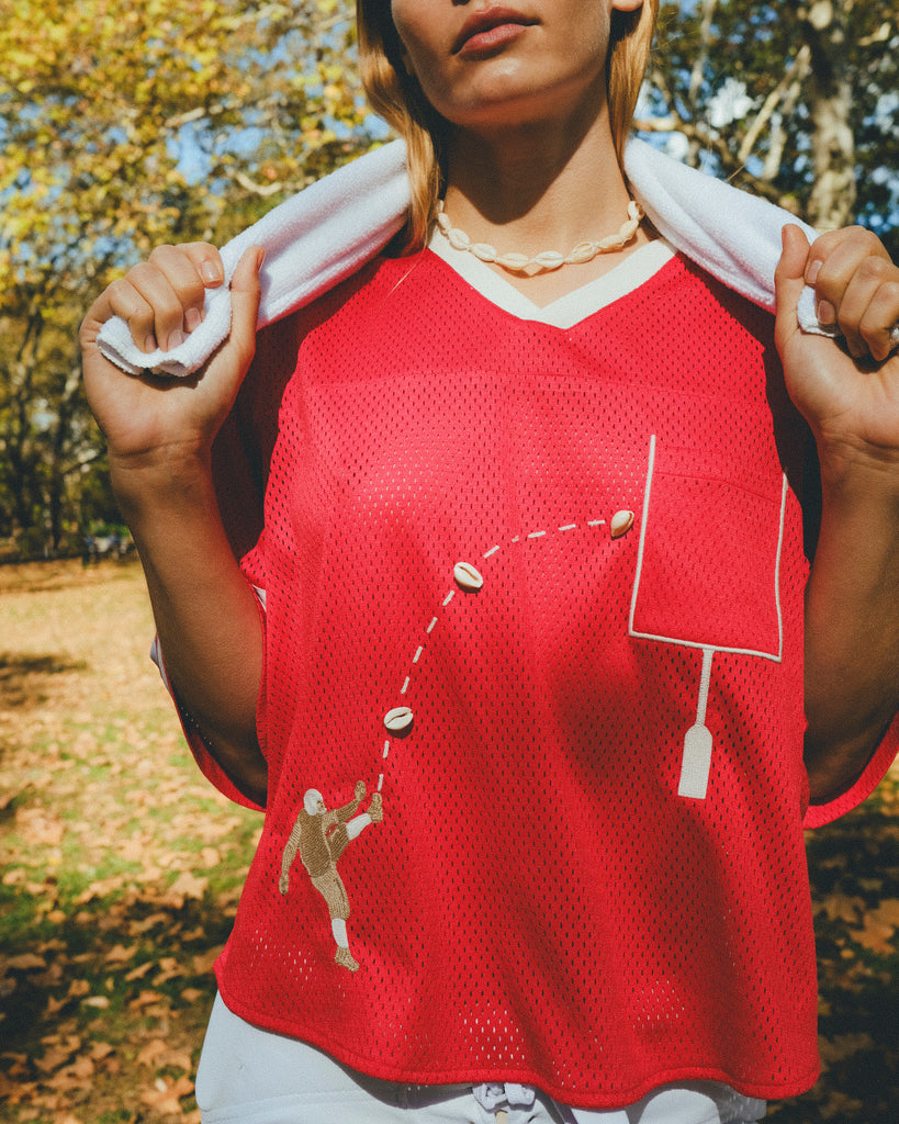 Close-up of a red mesh football-style top with cowrie shells forming a football graphic and an outlined pocket, worn over a white shirt with a towel draped around the neck.