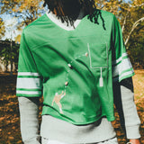 A man wearing a green mesh football-style crop top with cowrie shells forming a football graphic, layered over a gray long-sleeve shirt and paired with gray athletic shorts, holding shoulder pads in one hand.