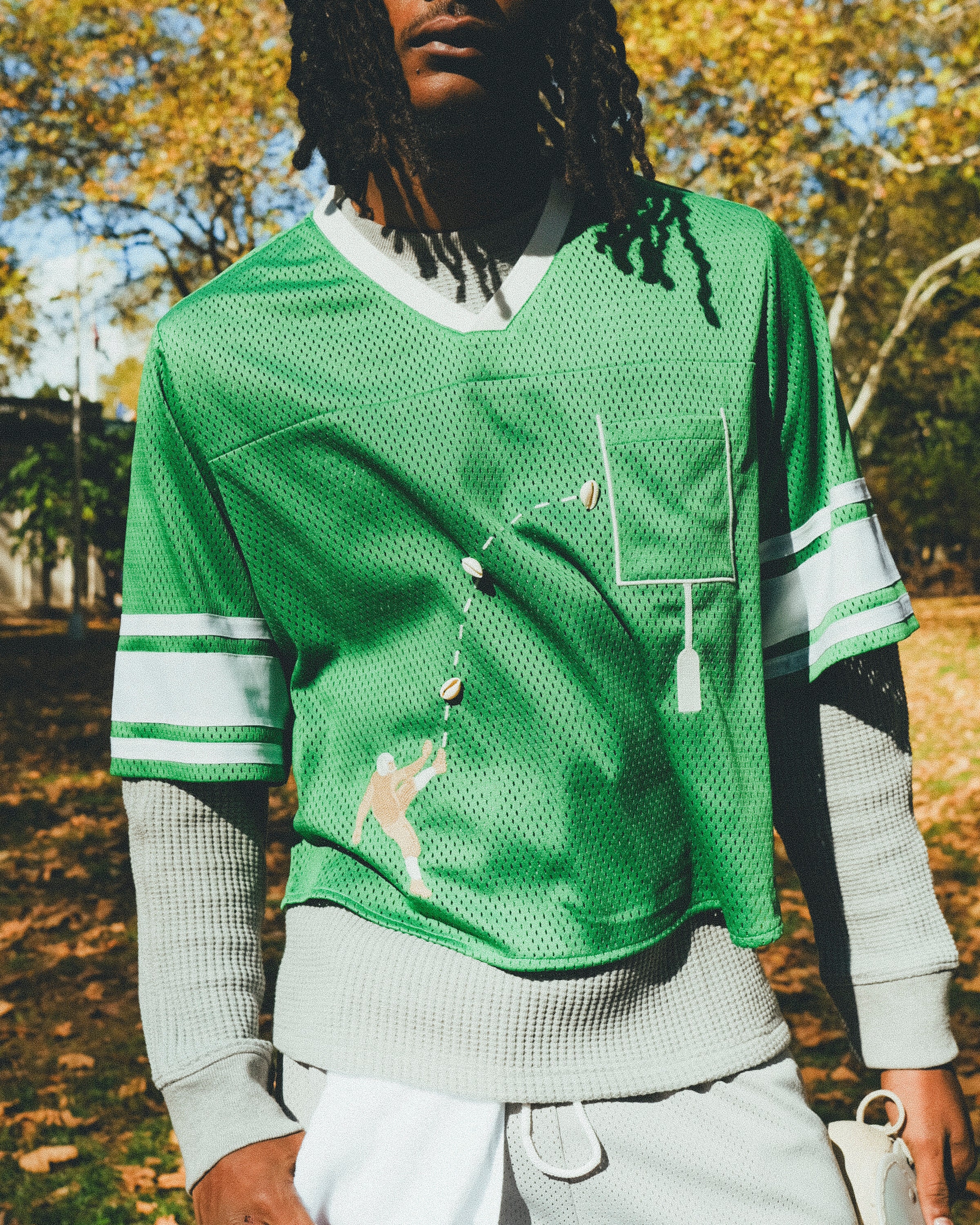 A man wearing a green mesh football-style crop top with cowrie shells forming a football graphic, layered over a gray long-sleeve shirt and paired with gray athletic shorts, holding shoulder pads in one hand.