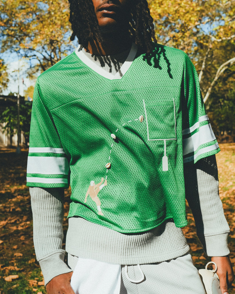 A man wearing a green mesh football-style crop top with cowrie shells forming a football graphic, layered over a gray long-sleeve shirt and paired with gray athletic shorts, holding shoulder pads in one hand.