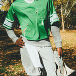 A man wearing a green mesh football-style crop top with cowrie shells forming a football graphic, layered over a gray long-sleeve shirt and paired with gray athletic shorts, holding shoulder pads in one hand.