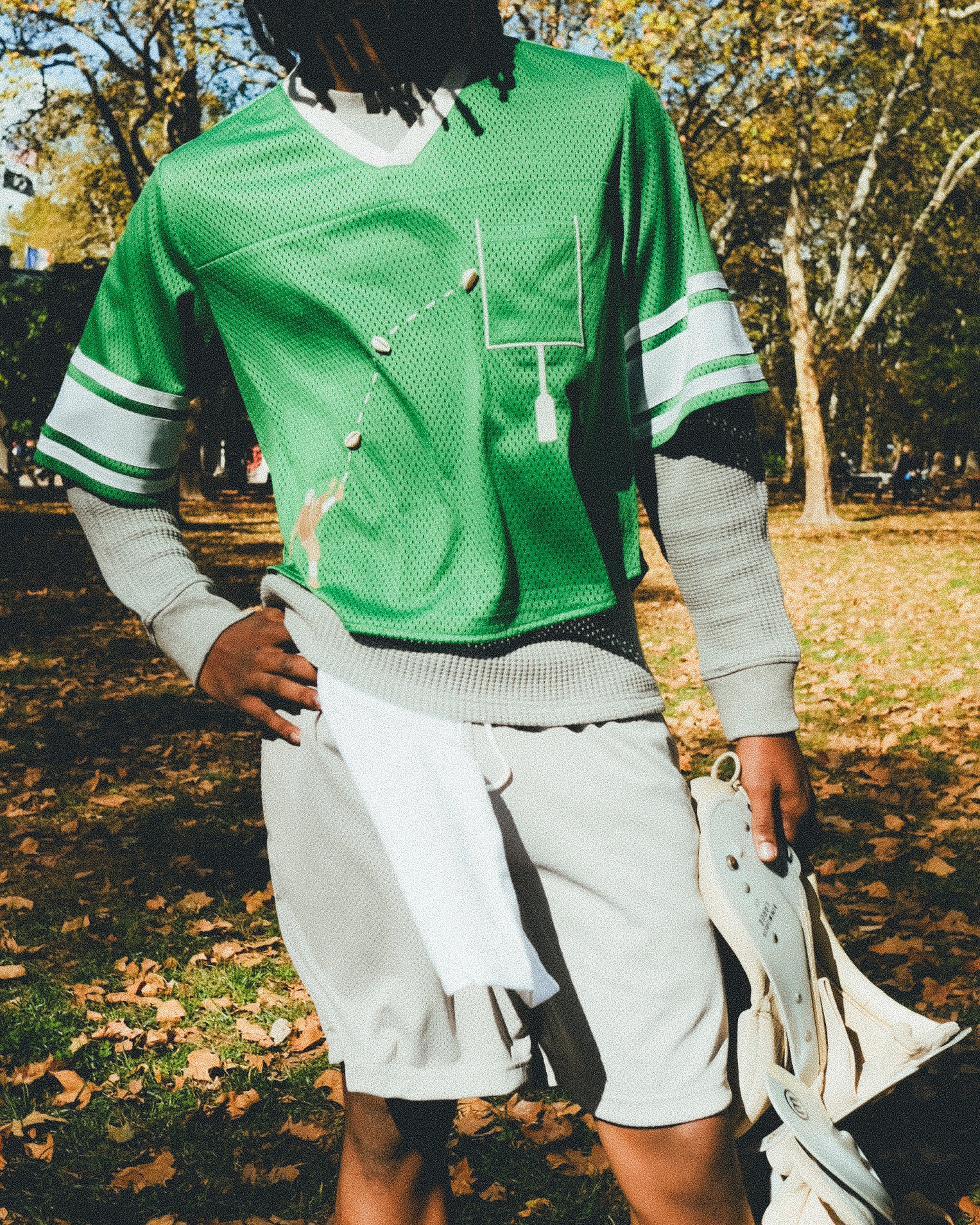 A man wearing a green mesh football-style crop top with cowrie shells forming a football graphic, layered over a gray long-sleeve shirt and paired with gray athletic shorts, holding shoulder pads in one hand.