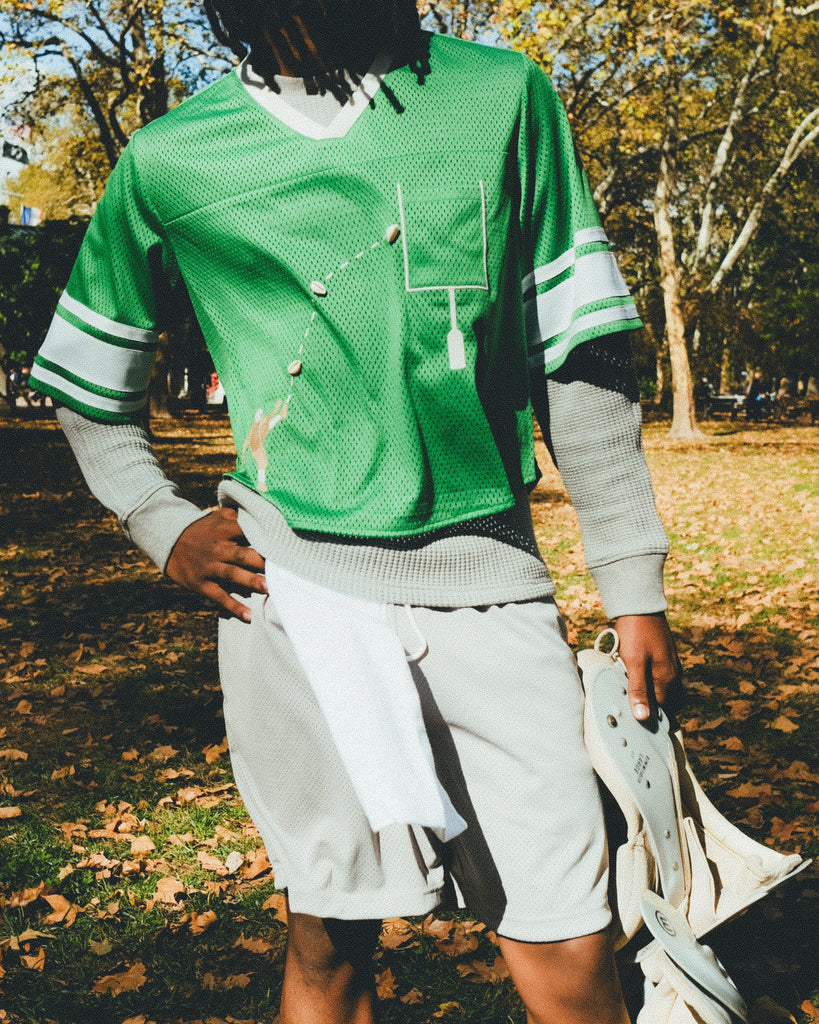 A man wearing a green mesh football-style crop top with cowrie shells forming a football graphic, layered over a gray long-sleeve shirt and paired with gray athletic shorts, holding shoulder pads in one hand.
