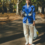 Female model wearing a blue zip-up knit sweater with a fish design, walking outdoors on a tree-lined path while holding a newspaper and a shopping bag.