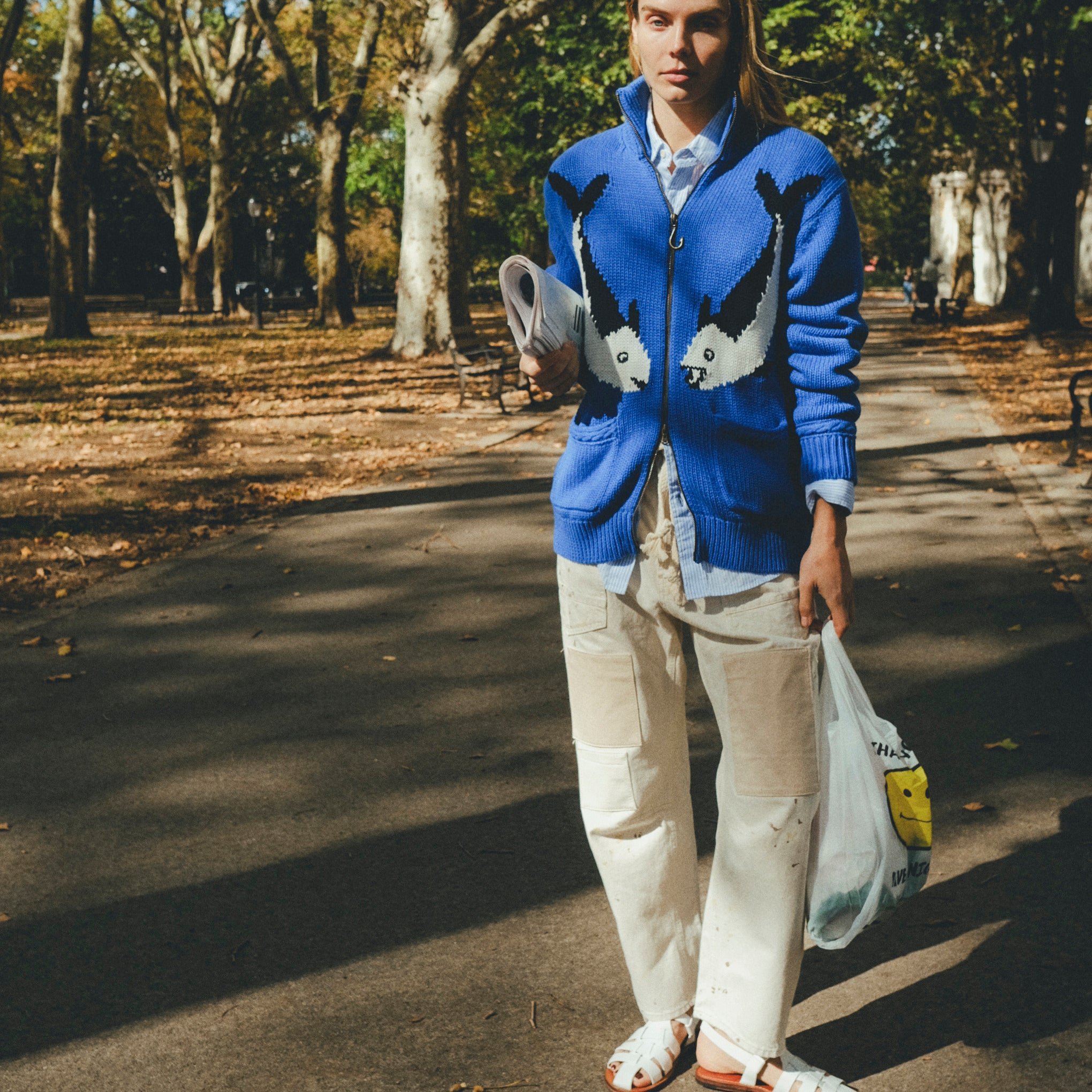Female model wearing a blue zip-up knit sweater with a fish design, walking outdoors on a tree-lined path while holding a newspaper and a shopping bag.