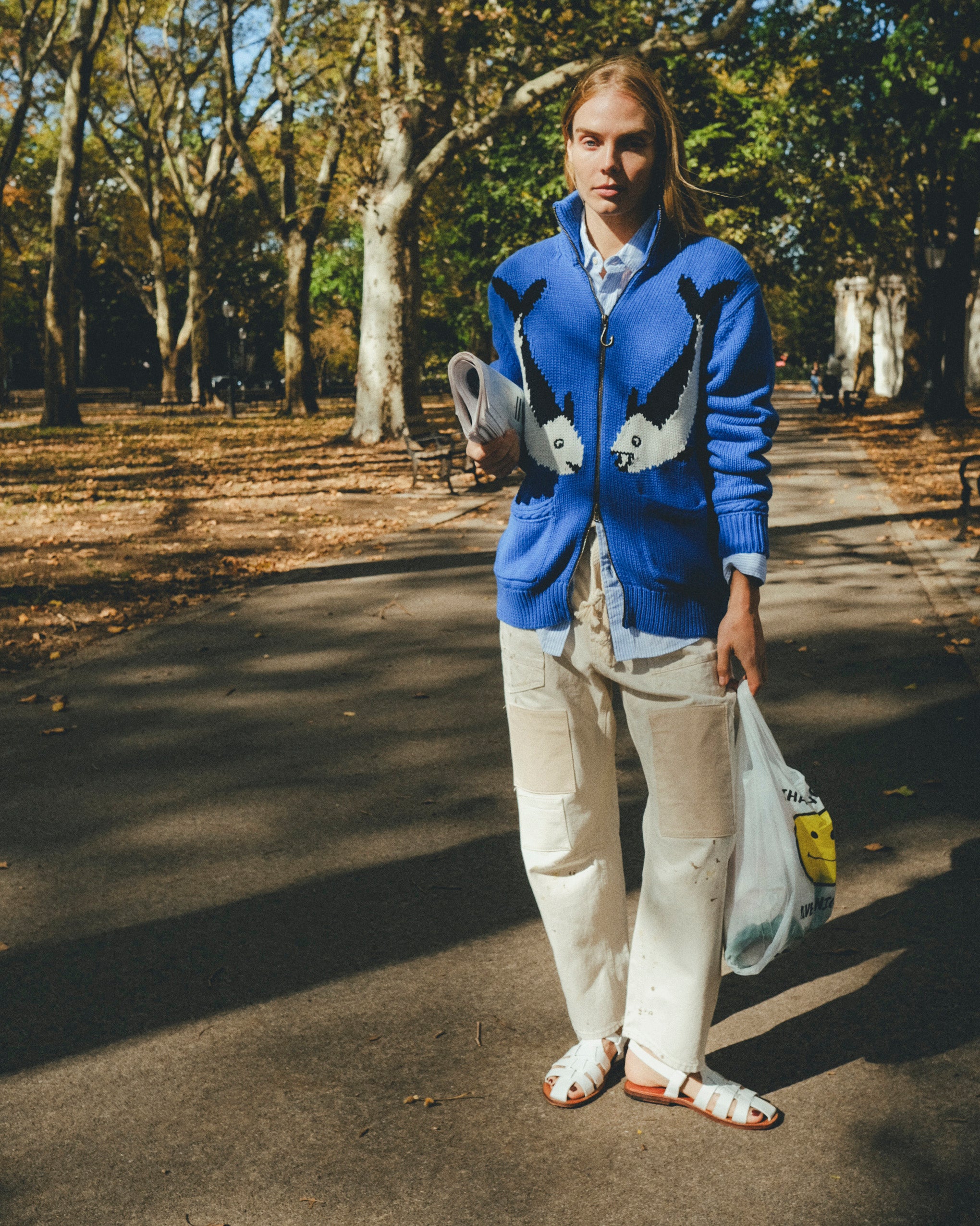 Female model wearing a blue zip-up knit sweater with a fish design, walking outdoors on a tree-lined path while holding a newspaper and a shopping bag.