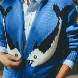Close-up of a blue zip-up knit sweater featuring a large fish design across the front, worn over a striped button-down shirt.