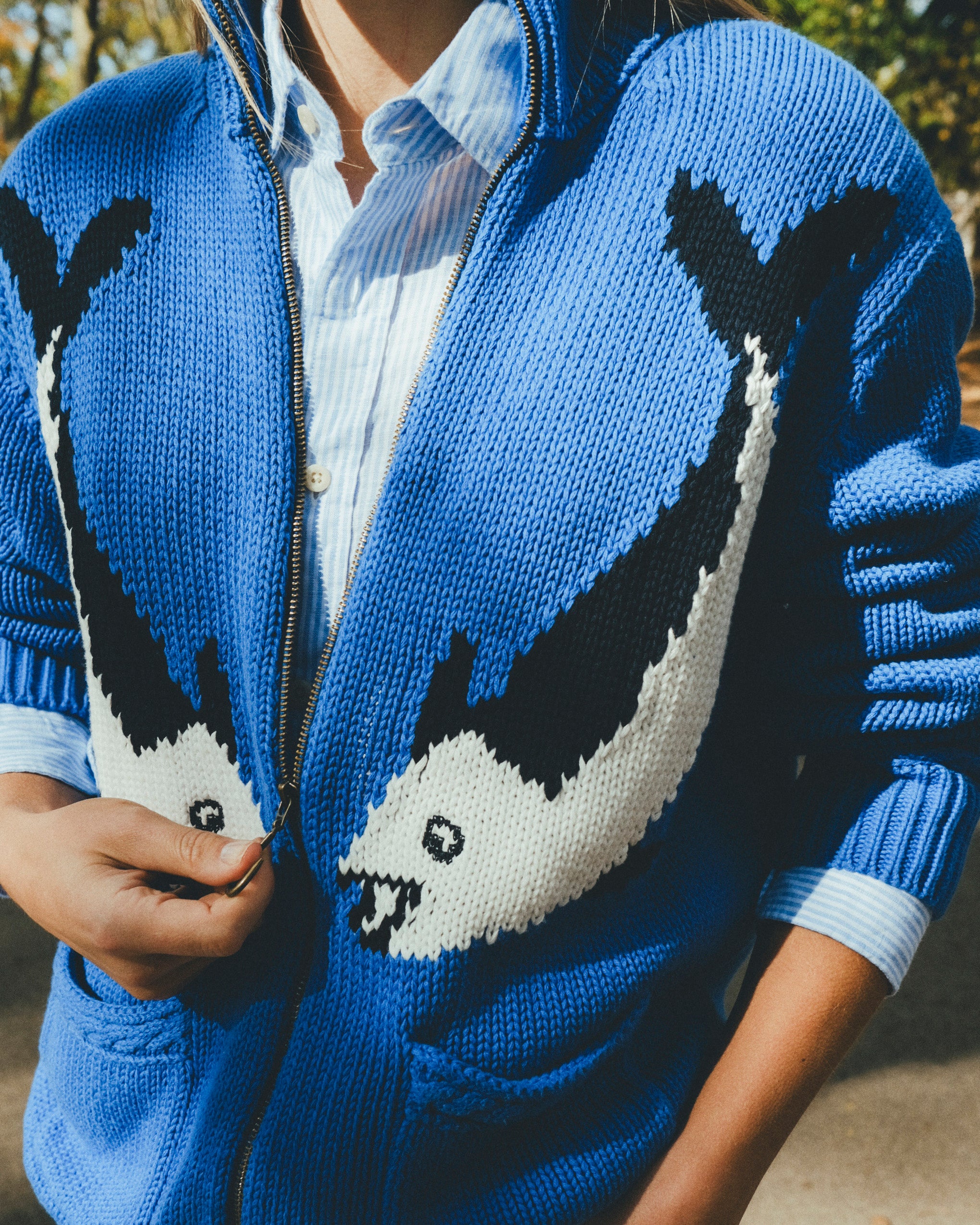 Close-up of a blue zip-up knit sweater featuring a large fish design across the front, worn over a striped button-down shirt.