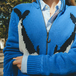 Close-up of a blue zip-up knit sweater featuring a fish design across the front, worn over a striped button-down shirt.