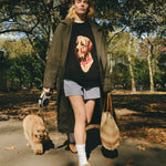Woman wearing a black knit sweater with an intarsia dog portrait on the front and a red collar detail, walking a dog through the park.