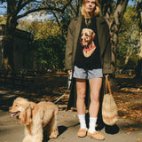 Woman wearing a black knit sweater with an intarsia dog portrait on the front and a red collar detail, walking a dog through the park.